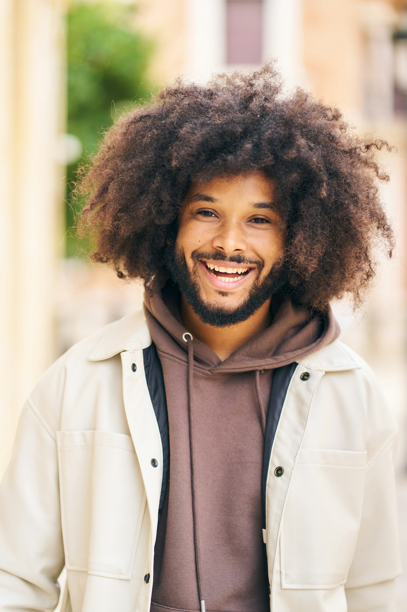 A man with curly hair is smiling and wearing a brown hoodie